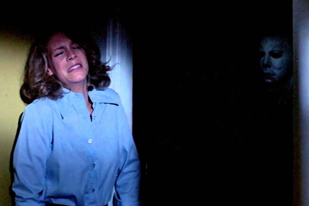 Laurie Strode hiding with her back against a wall, lit up by the moonlight shining through a window. Michael Myers a.k.a. The Shape is lurking in the darkness behind her. His body dark but his pale white, emotionless mask is lit up by the moonlight behind her.