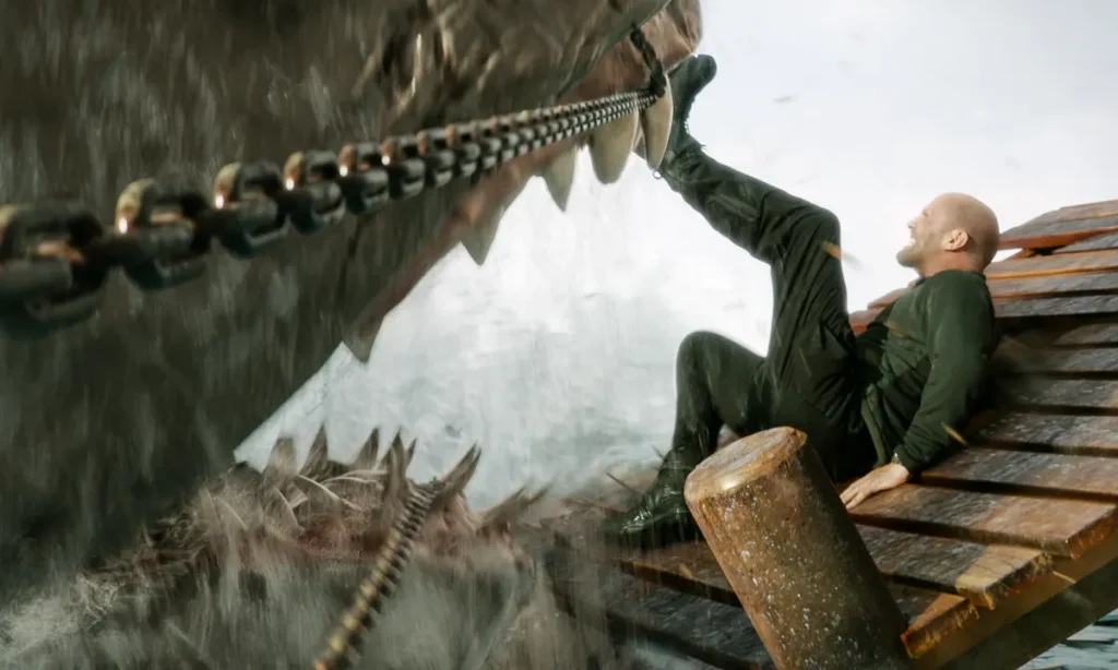 The Meg attempting to eat Jonas, played by Jason Statham, who is leaning on a tipped and broken boardwalk. Jonas is using his foot to kick the upper teeth of the Meg, which is also being restrained slightly by a chain across its face.