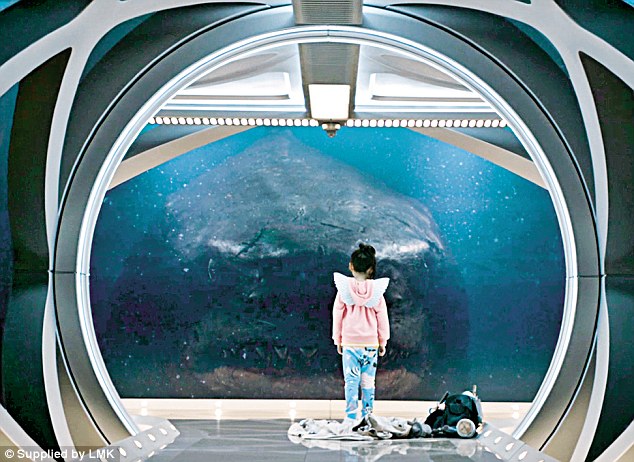 Small girl standing in front of a deep, dark underwater window, and looking back at her is a prehistoric megalodon shark with menacing eyes and sharpened teeth.