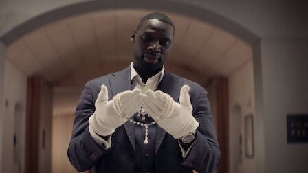 Assane, suited up, holding the Queen's necklace in the delicate grasp of his white gloves.