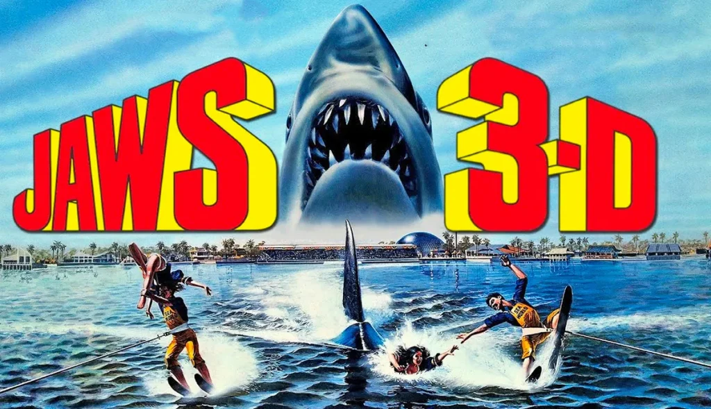 The Jaws 3D Poster, 1983. A townscape, with a large shark head emerging from water behind it. In front, people are under attack and water skiing away from a shark. In large 3D writing, in bright red and yellow, 'JAWS 3-D' is branded across the poster.